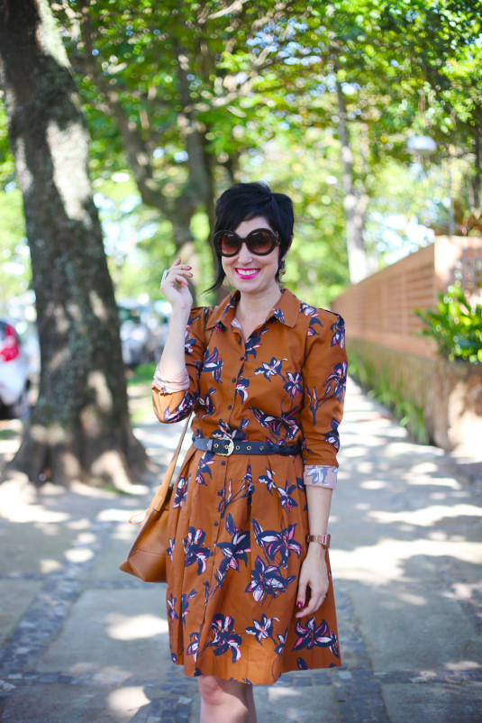 vestido marrom claro com flores azuis e rosas grandes, com gola tipo camisa e mangas compridas, saia rodada na altura dos joelhos, cinto preto trançado na cintura, oxford rosé e bolsa saco caramelo